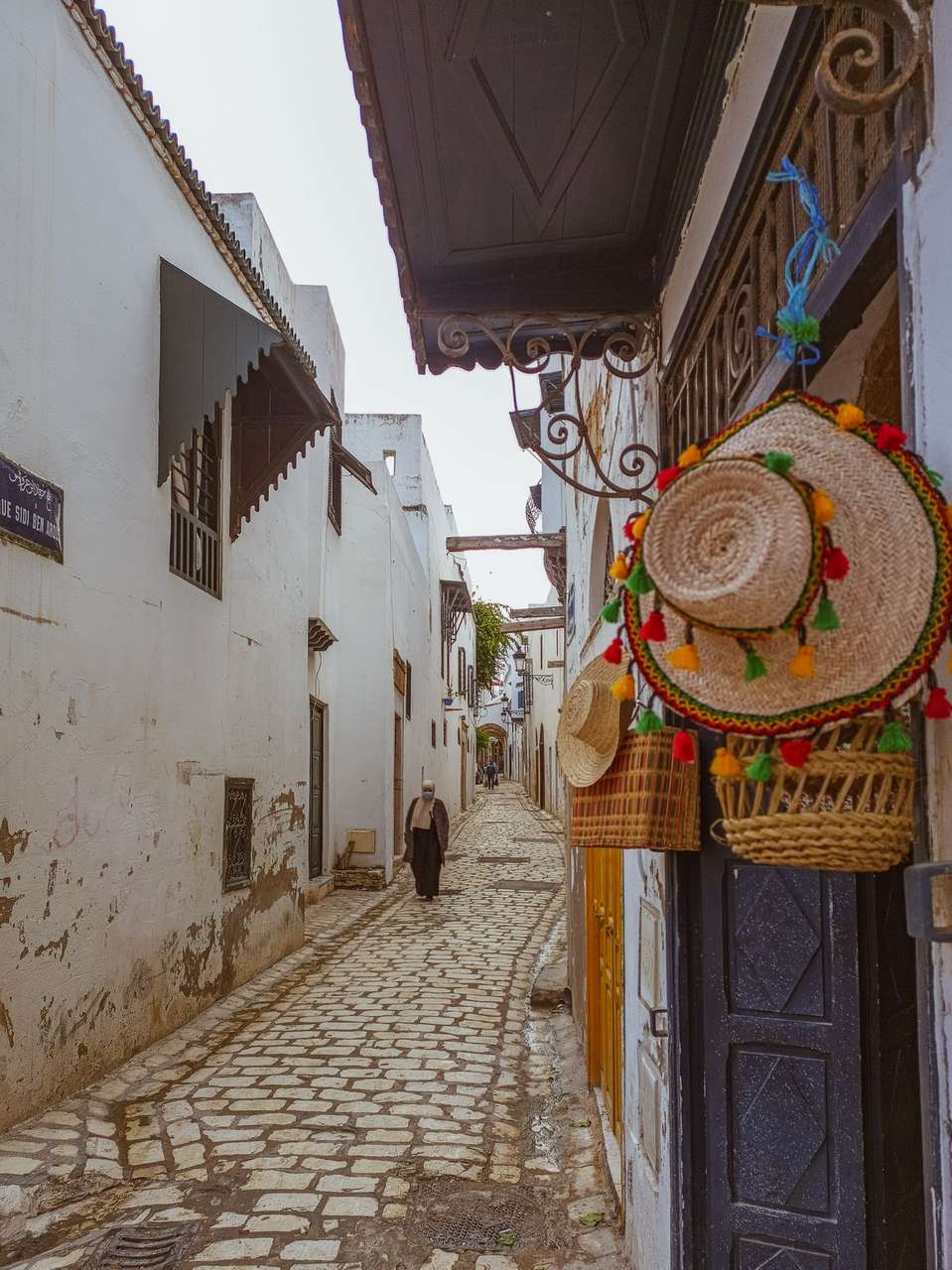 Narrow cobblestone alley in the Tunis medina with white walls and traditional craft shop selling straw hats, Tunisia